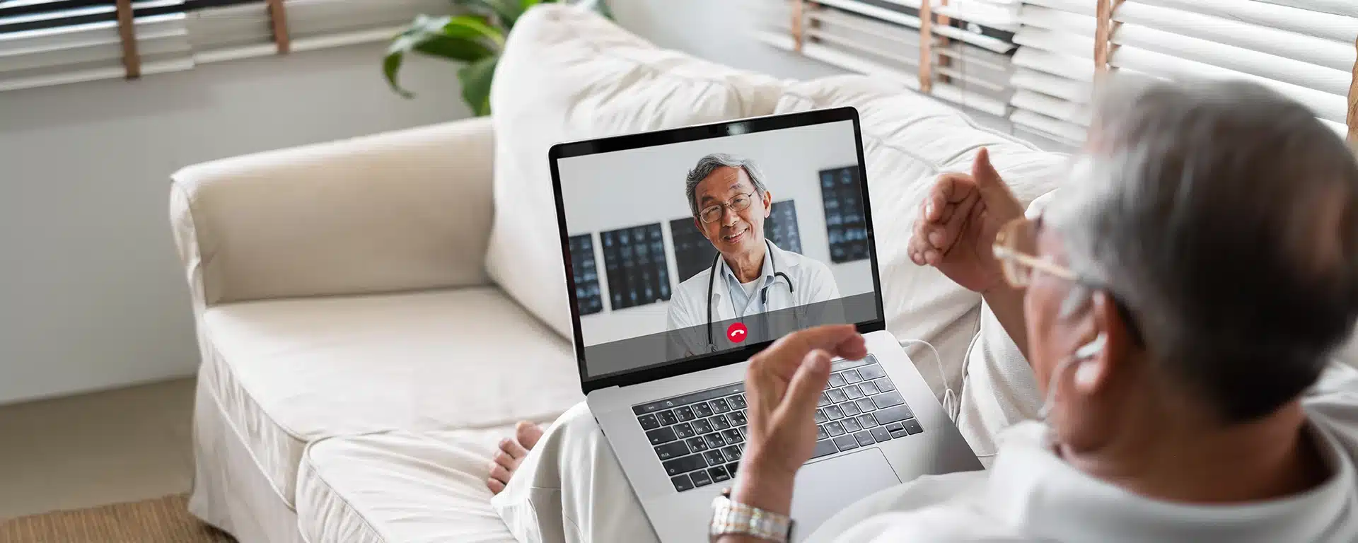 A senior man sits on a couch while conversing with his doctor through a tele-health service.